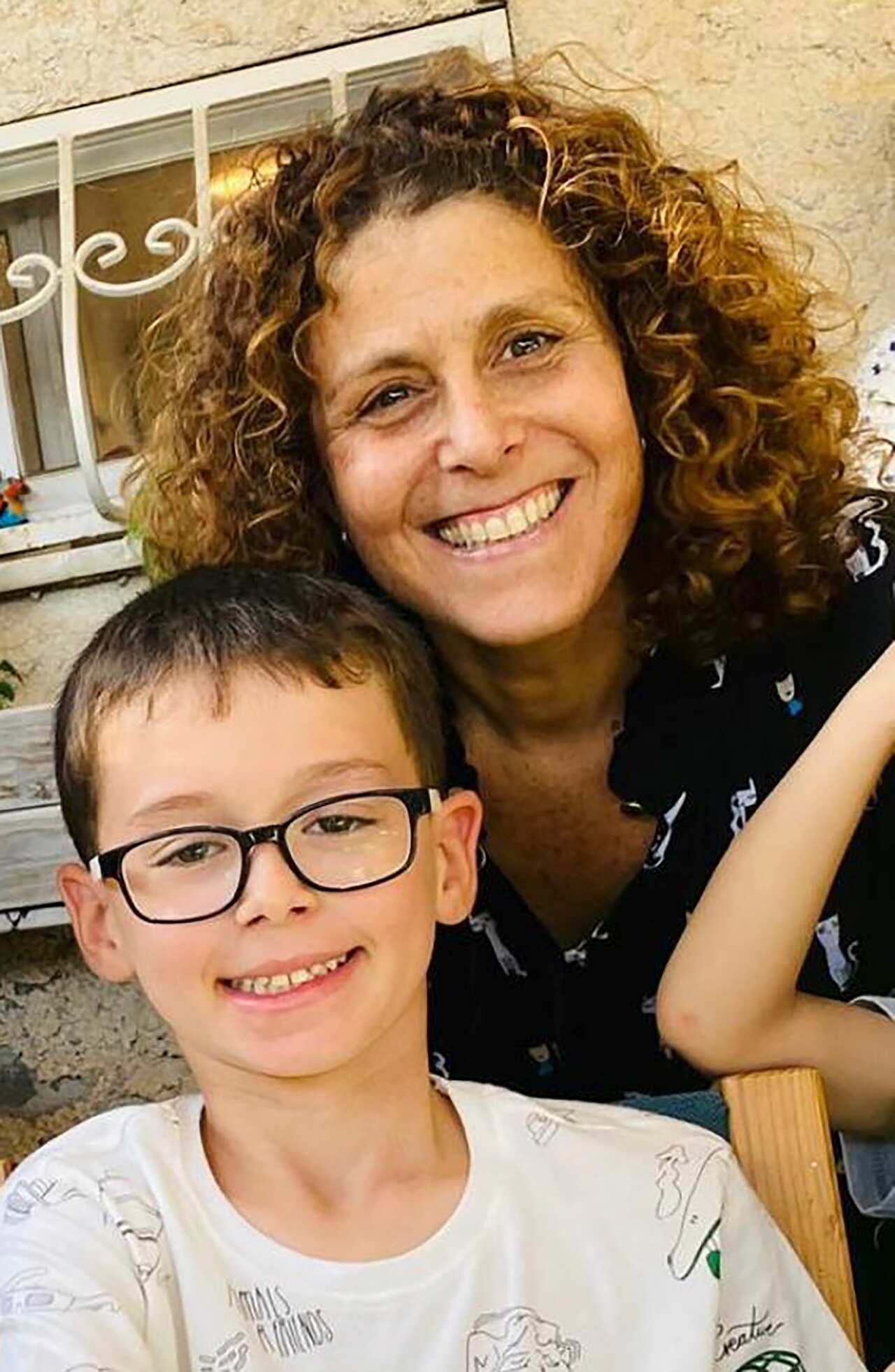 A woman with curly hair smiles with her cheek pressed up against the head of a young boy with short hair and glasses. 