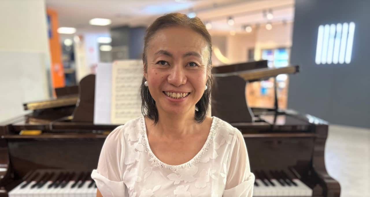 A woman in a white shirt sits in front of a grand piano smiling at camera.