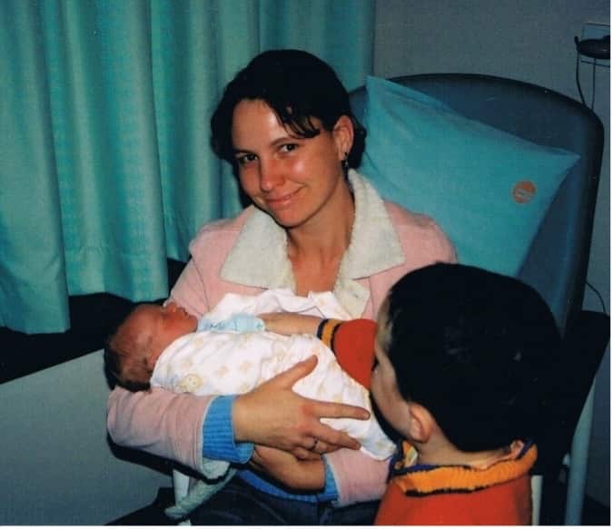 A smiling woman with short brown hair holds a newborn baby, while another young child looks on.