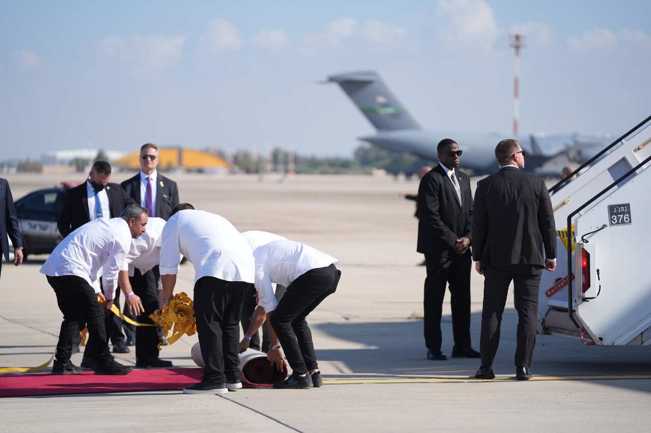 A group of workers roll out a red carpet beside a plane, where two agents in black suits wait at the bottom of the stairs.