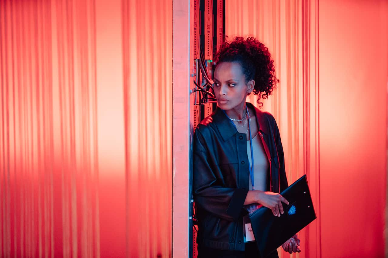 A woman holding a folder stands in front of a bright orange wall, looking sideways.