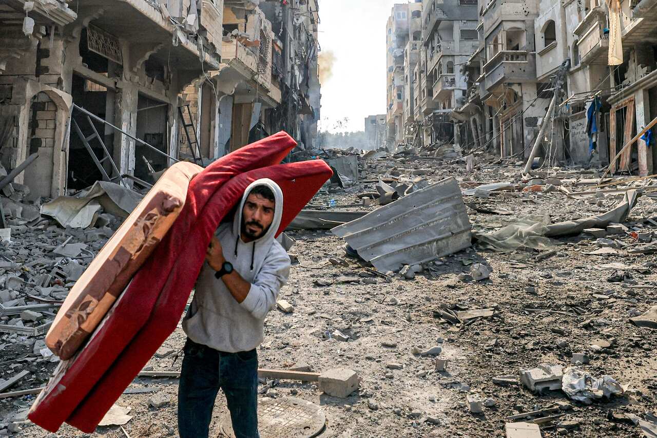 Man in grey hoodie and black pants walks through rubble, carrying three thin mattresses. Damaged buildings surround him. 