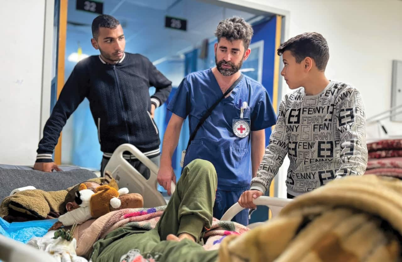 A bearded male nurse in a blue uniform stands next to a patient on a hospital bed with two young men looking at him as they stand on either side.