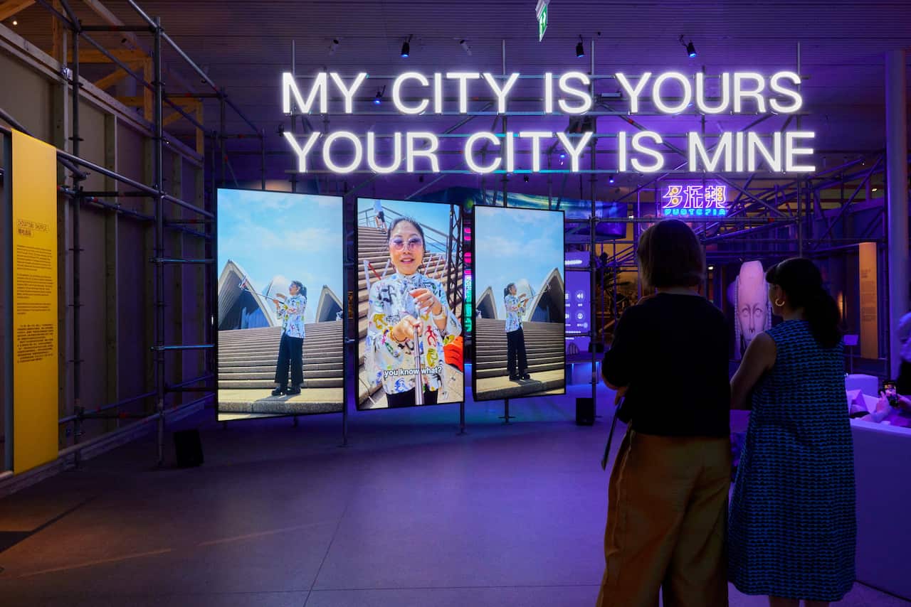The inside of an exhibition shows a three-panel video installation and neon signage that reads: "My city is yours your city is mine."
