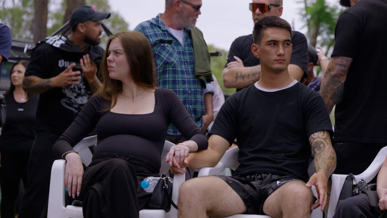 A man and a pregnant woman are sitting in white plasic chairs outside and holding hands. There are people standing and talking behind them