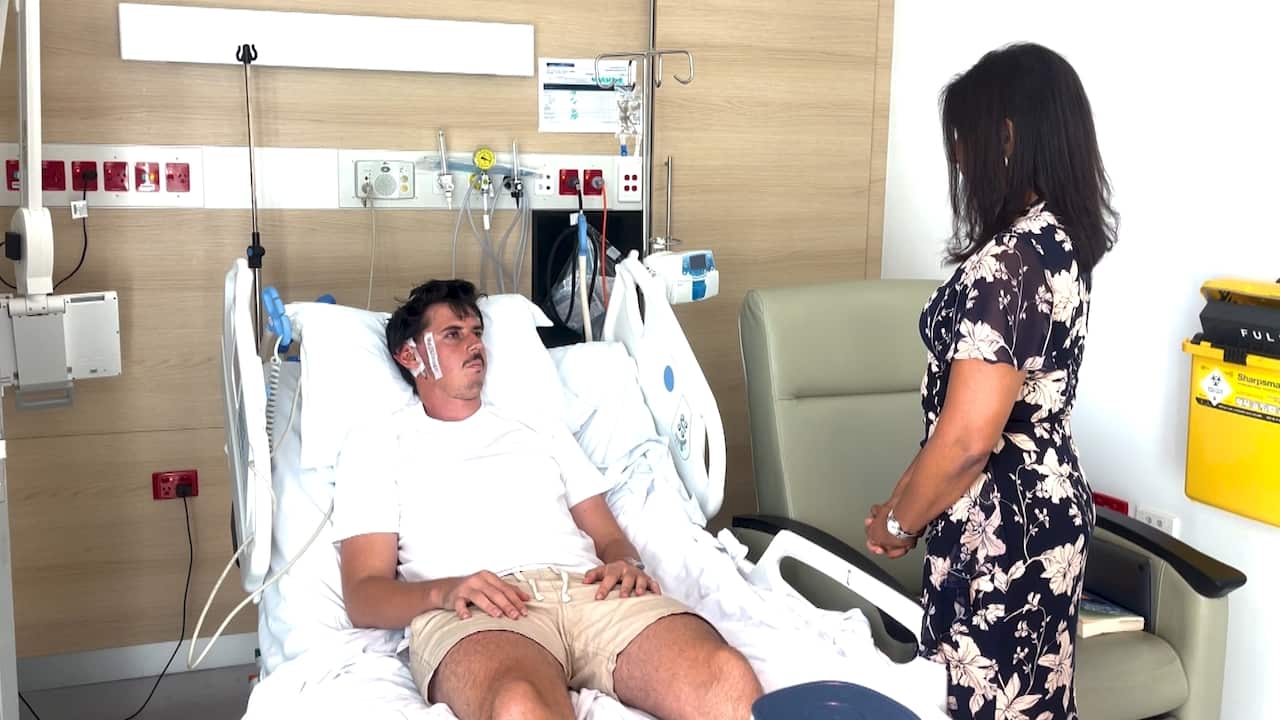 A man reclines at a 45 degree angle in a hospital bed. A woman in a floral dress stands above him.