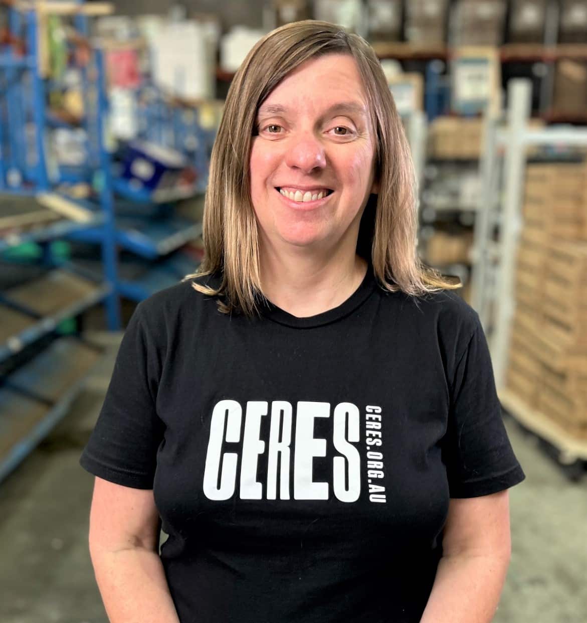 A woman in a black t-shirt facing camera and standing in a warehouse. 