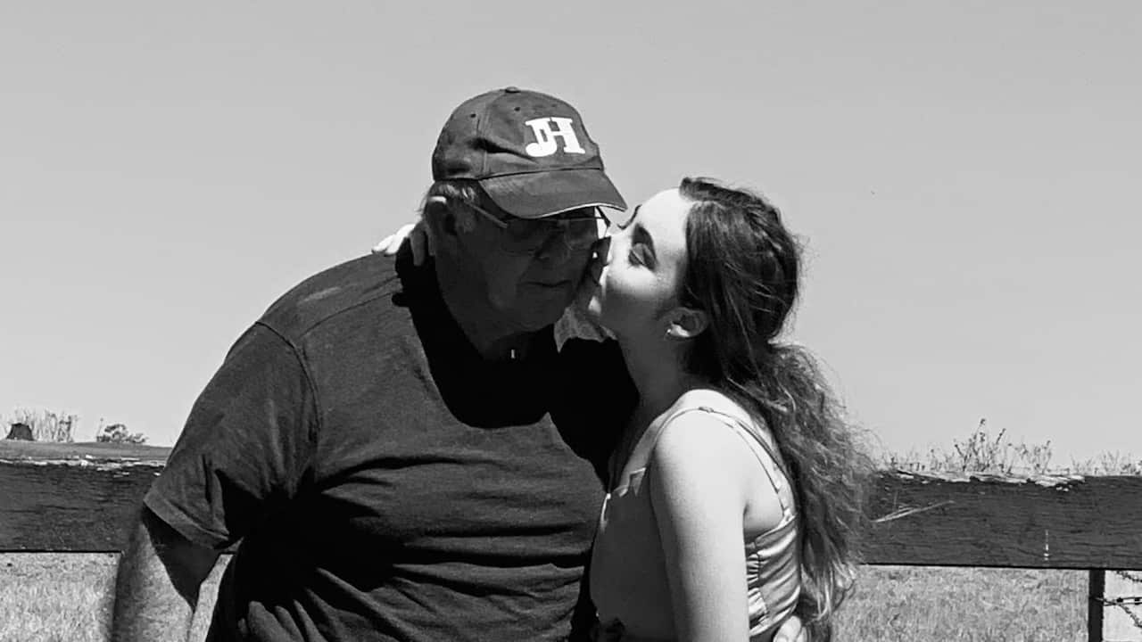 a black and white photo of a teenage girl kissing an old man (her father) on the cheek in front of a paddock fence