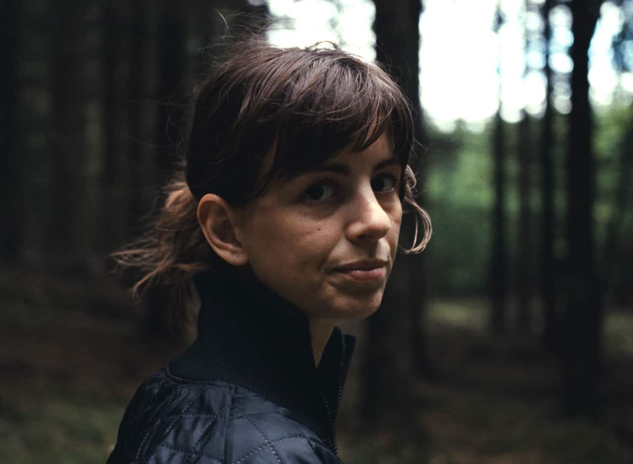 A woman is seen in a head and shoulders image. She stands in a wood, turning to look at the camera. 