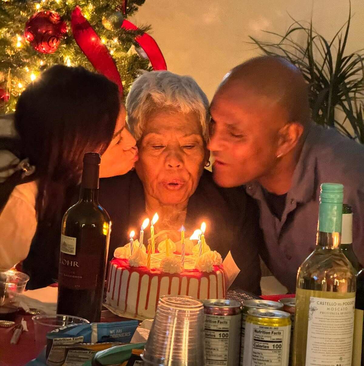 A younger man and woman lean in to kiss the cheeks of an older Vietnamese woman who is blowing out the candles of a cake. 