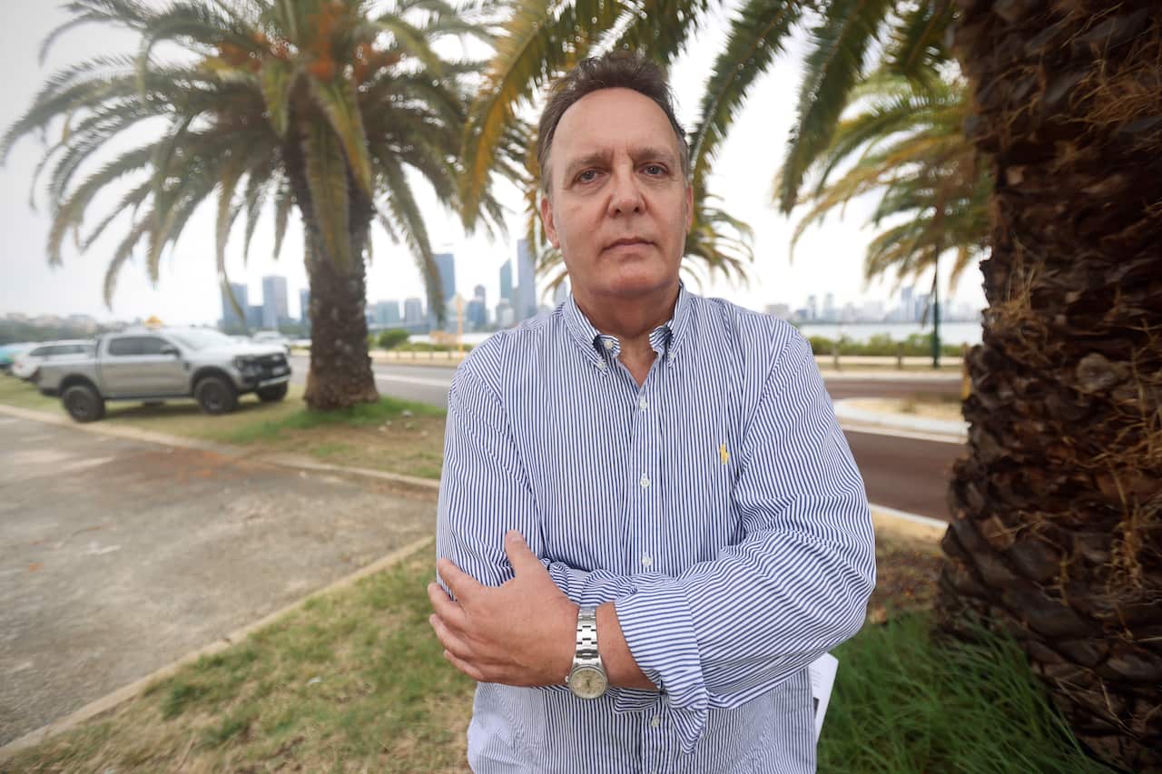 A middle-aged man looking at the camera, posing in front of a waterfront.
