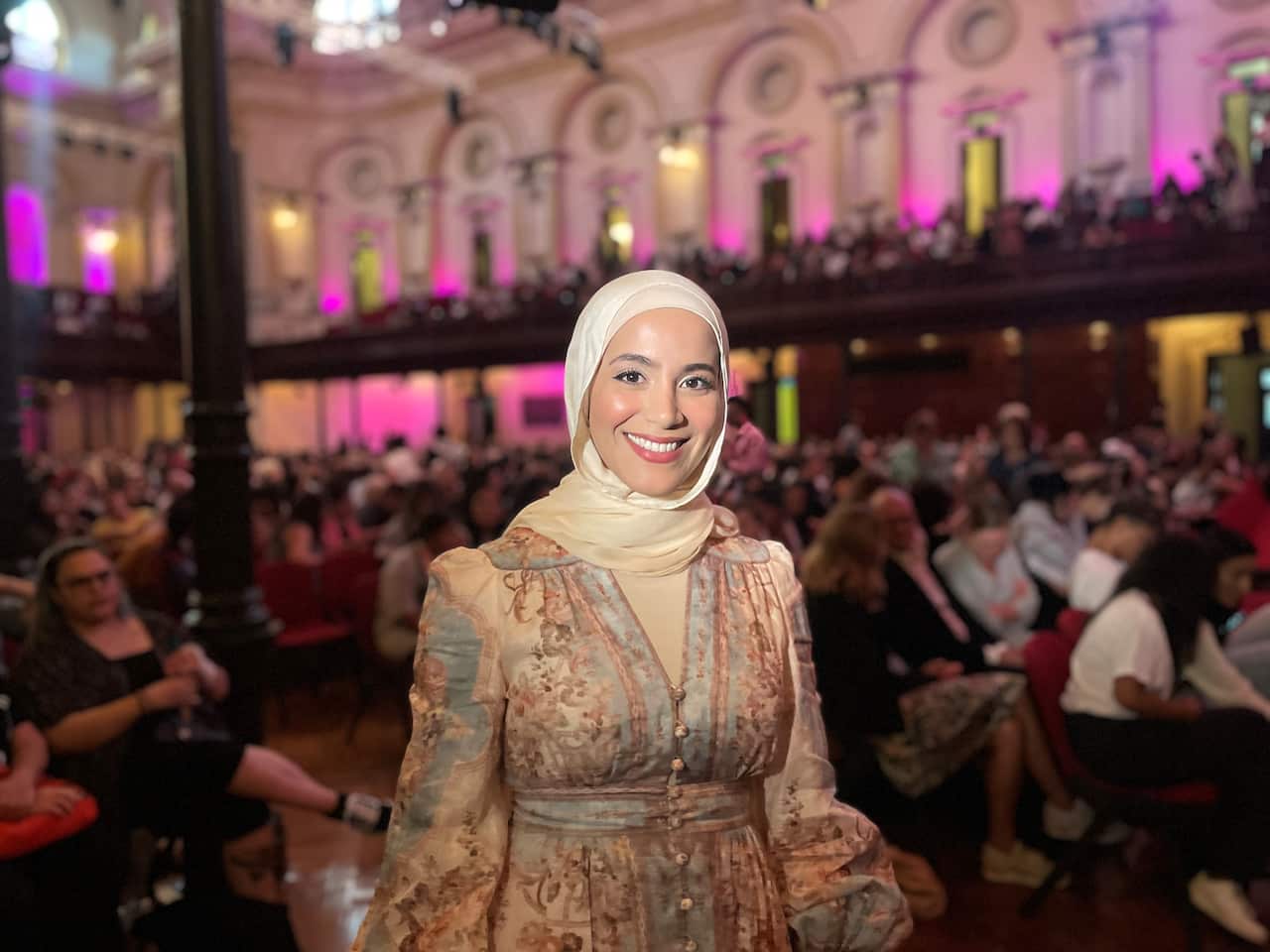 A woman in a cream hijab smiles in a floral dress behind a crowd of people in a hall. 