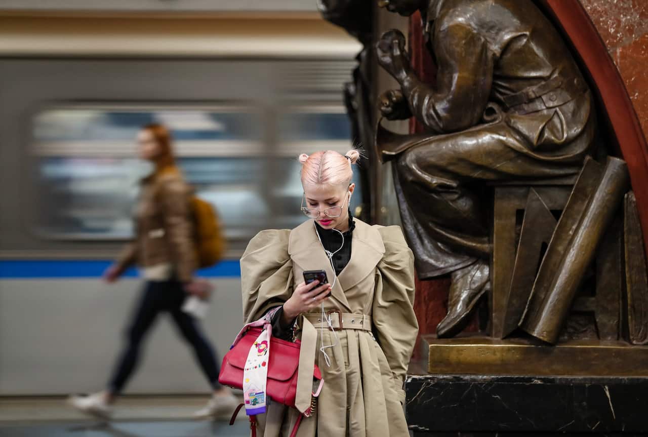 A woman in a tan coat using her phone next to a large bronze statue in a bustling subway station in Moscow.