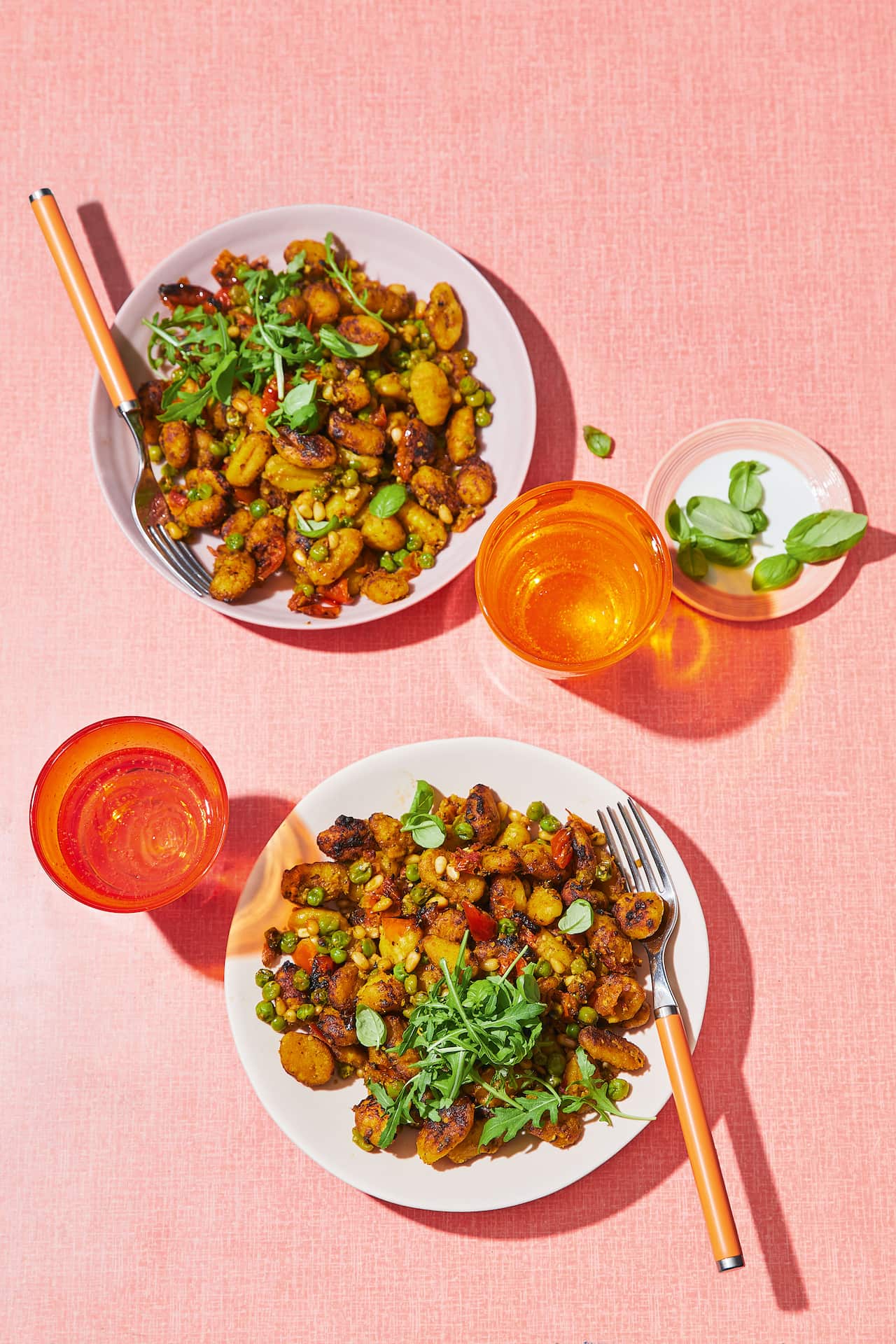 Seen from overhead, two plates with golden gnocchi sit on a blush-coloured tablecloth. Orange glasses sit alongside. 