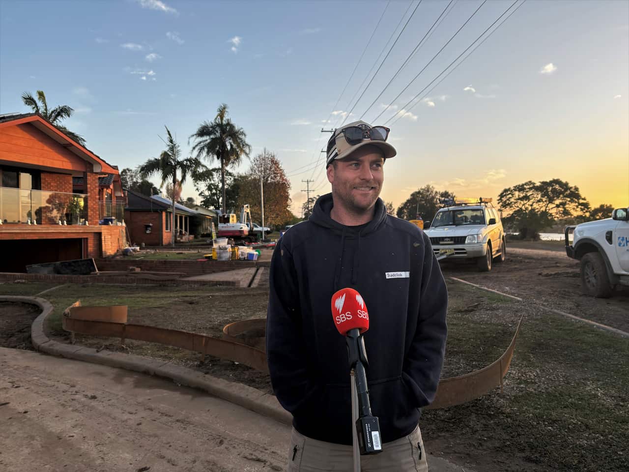 A young Caucasian man in his thirties smiling and looking off to the right. Wearing a beige cap with sunglasses on top of it, a black hoodie and light grey shorts. In the background is a street seen at sunset, machinery in the background, and signs of damage all around.
