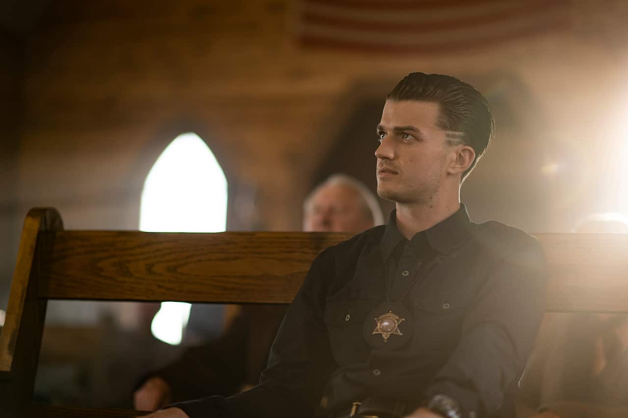 A young man in dark shirt, wearing a sheriff's badge, sits in a pew. 