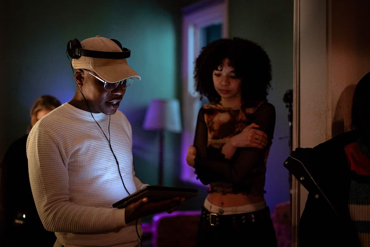 A man in a cap and headphones, and a young woman standing with crossed arms beside him, stand in a dark room, looking down at a tablet he is holdnig. 