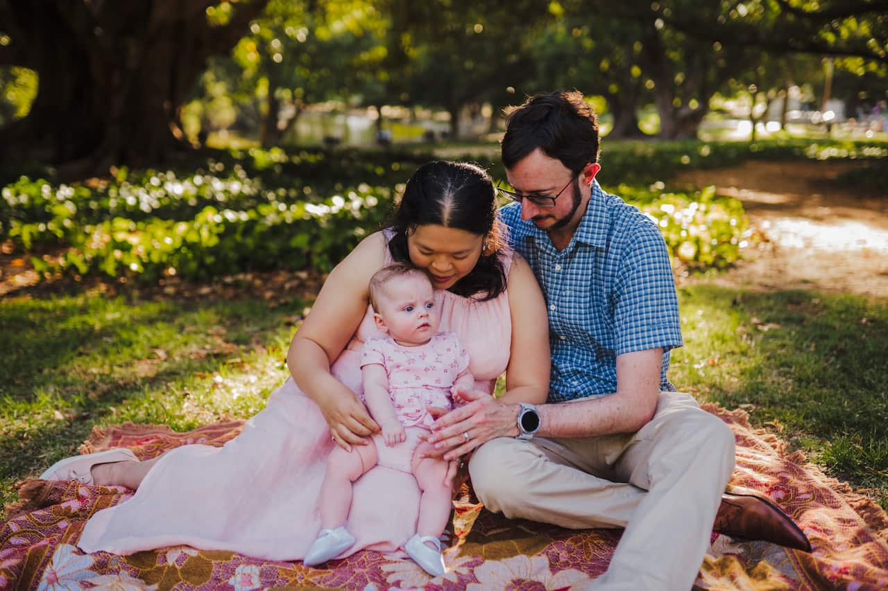 A mother and father sit with their daughter on a picnic blanket in the sunshine. The parents look down lovingly at their daughter.