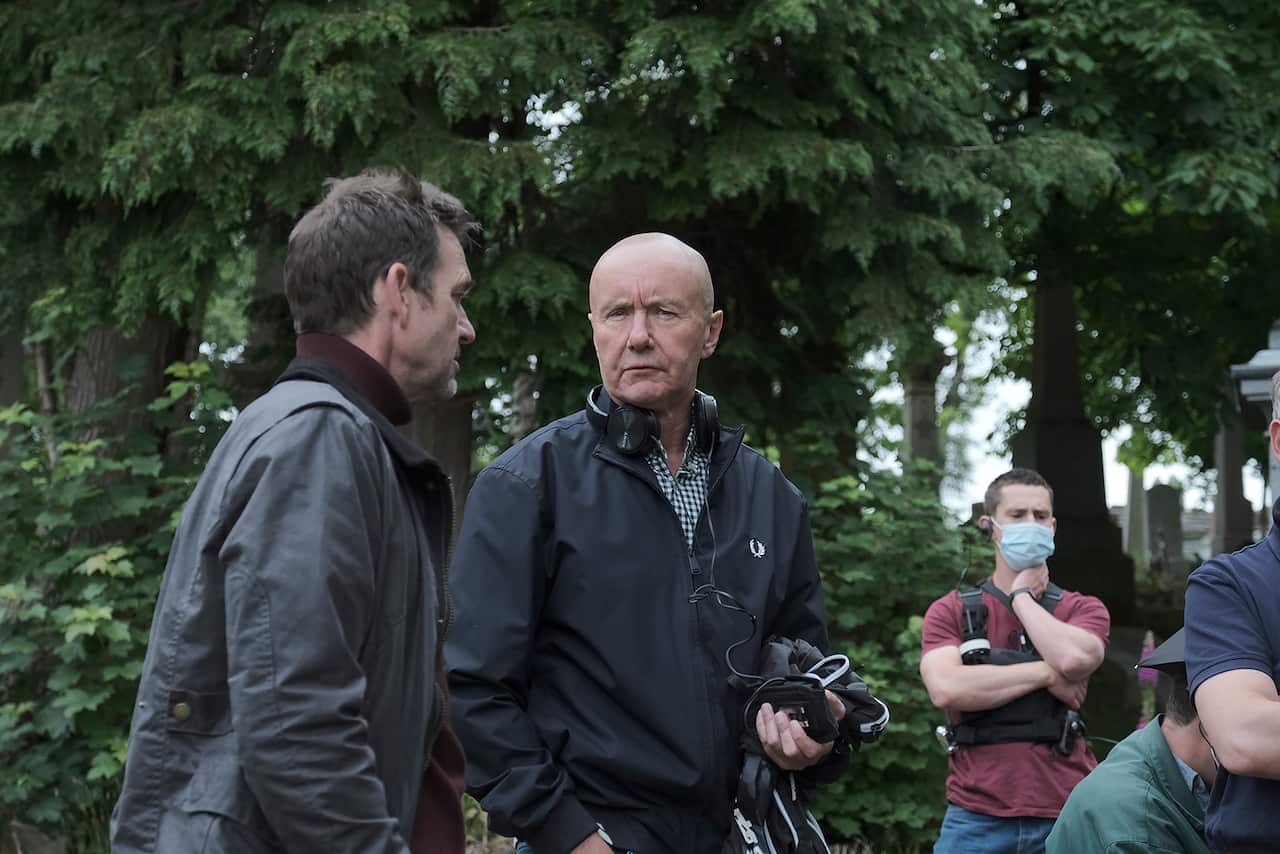 In a behind the scenes shot, a man stands in front of greenery, talking to a man who can be seen side-on. Members of the crew can be seen in the background.