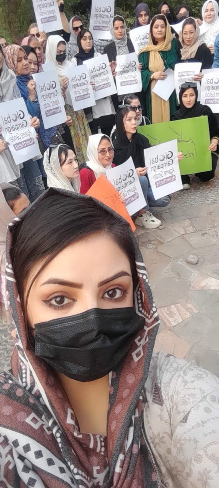 A woman in a headscarf and wearing a black mask over her mouth takes a selfie. Behind her are people holding protest placards