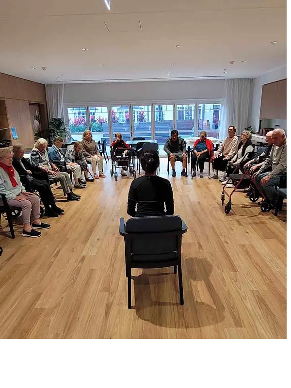 A woman in a chair in the centre of a circle, her back turned, surrounded by older people sitting in chairs.