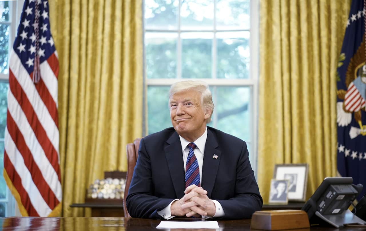 Donald Trump smiles while sitting in the Oval Office of the White House, with his hands crossed.