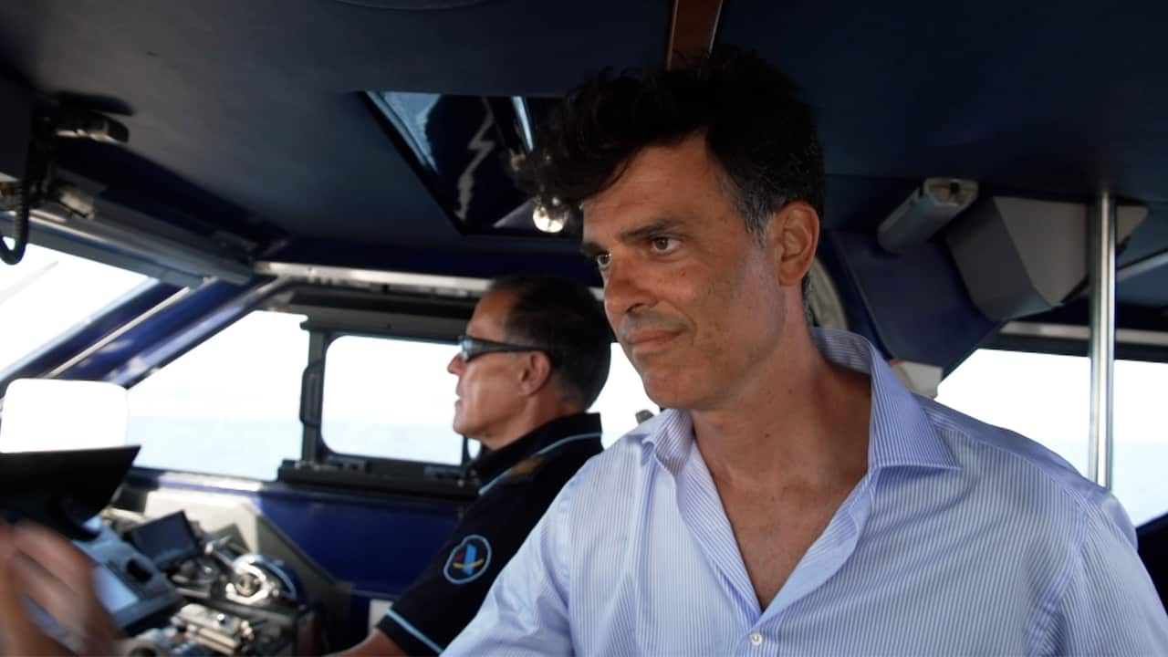 A tanned man wearing an unbuttoned blue shirt standing on a speedboat. Another man in a uniform and sunglasses is behind him at the dashboard and looking straight ahead 