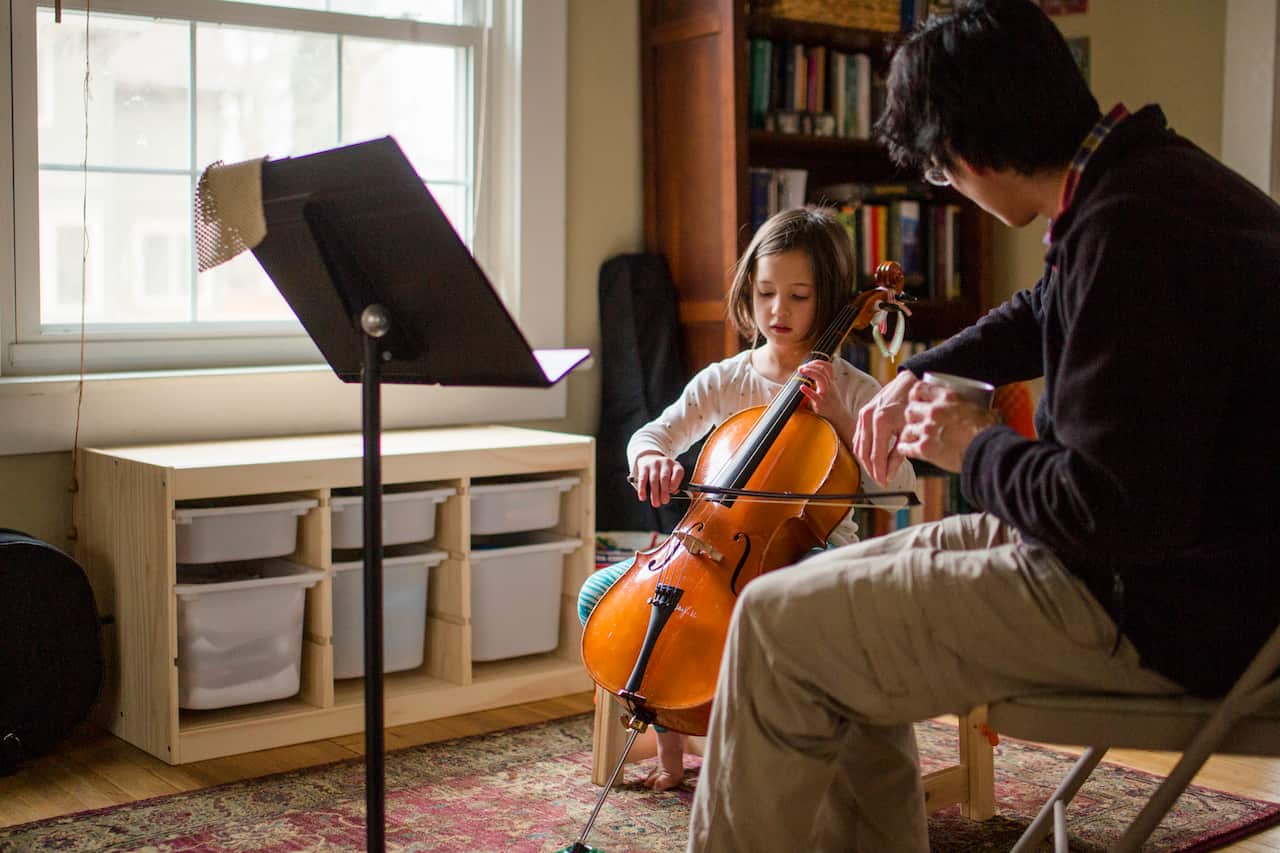 A father helps little girl practice cello at home by window - Australia Explained – Learning an instrument