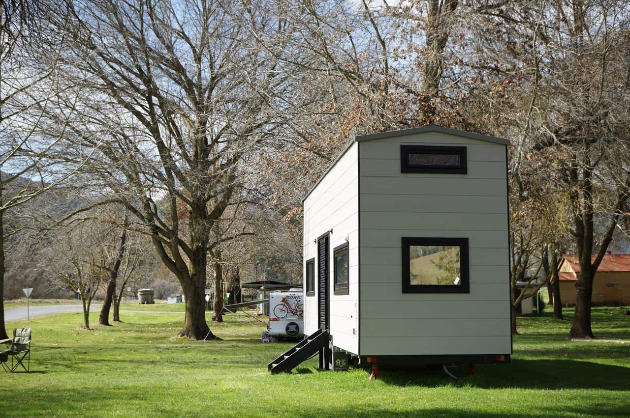 A white-clad tiny house on wheels is parked on lush green grass and is surrounded by trees. There's a small step ladder to enter inside the tiny house.