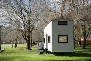 A white-clad tiny house on wheels is parked on lush green grass and is surrounded by trees. There's a small step ladder to enter inside the tiny house.