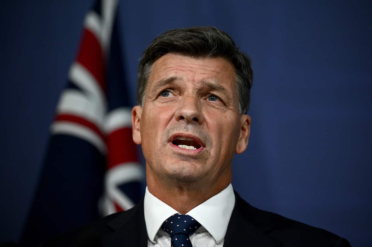 A man in a black suit is speaking in front of an Australian flag.