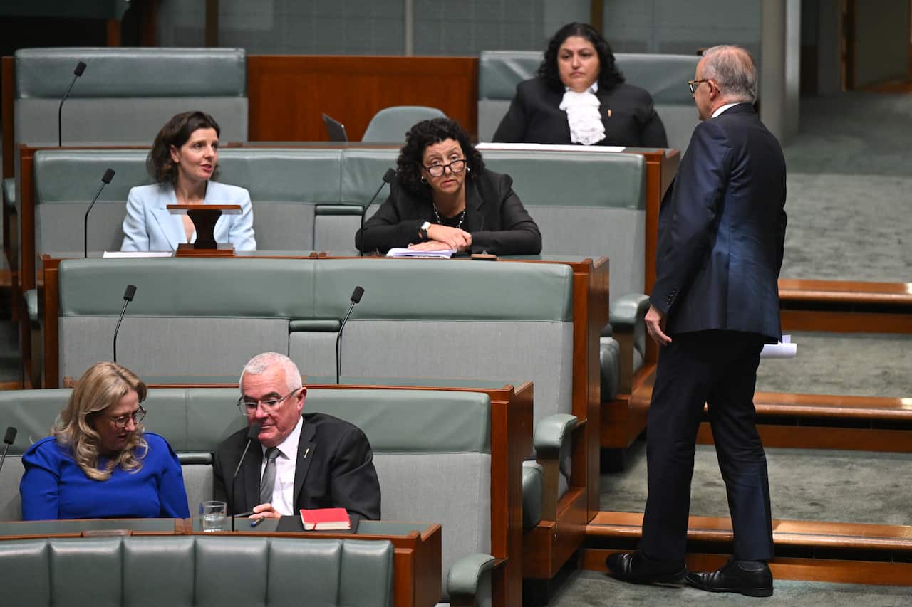 A man in a suit stands on the steps inside the House of Represenatives, talking to two women sitting down.