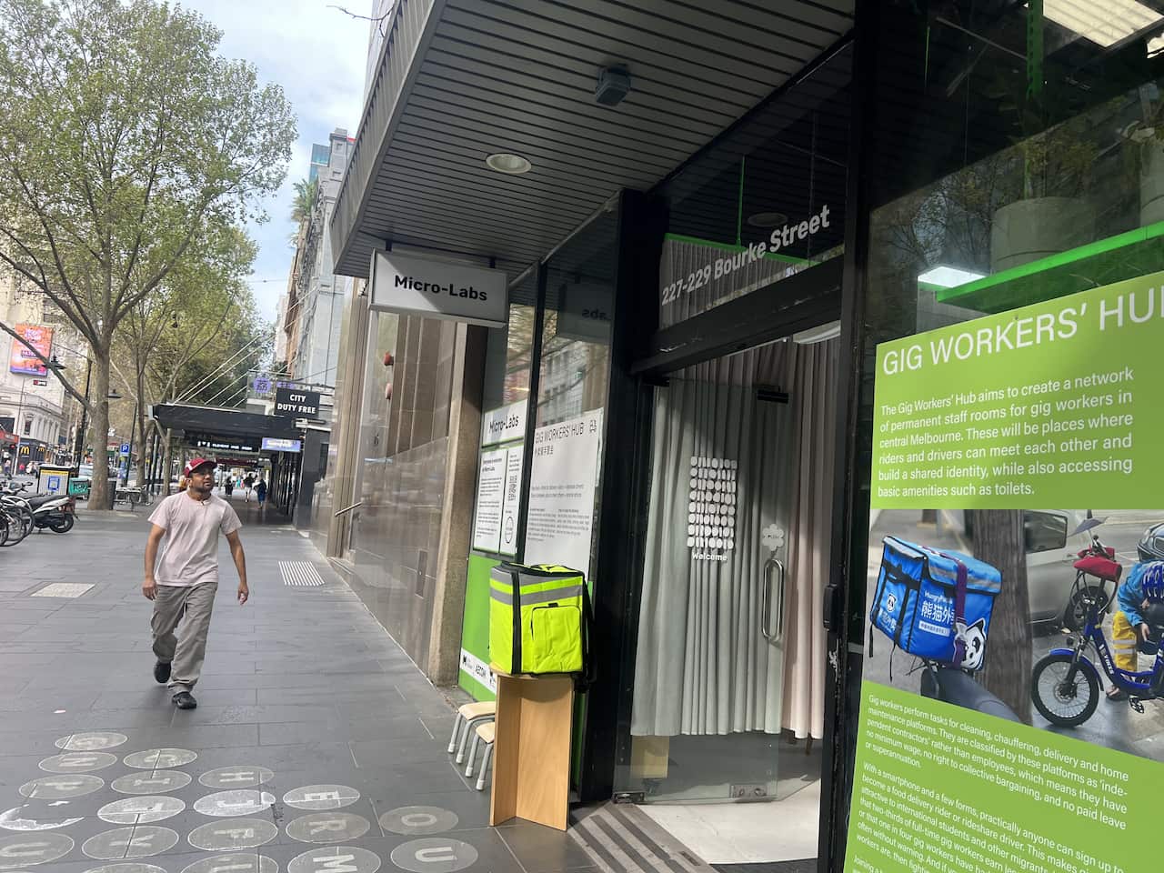 A man walks on along a city street near a shop front with a green billboard sign at the front that reads Gig Workers' Hub 