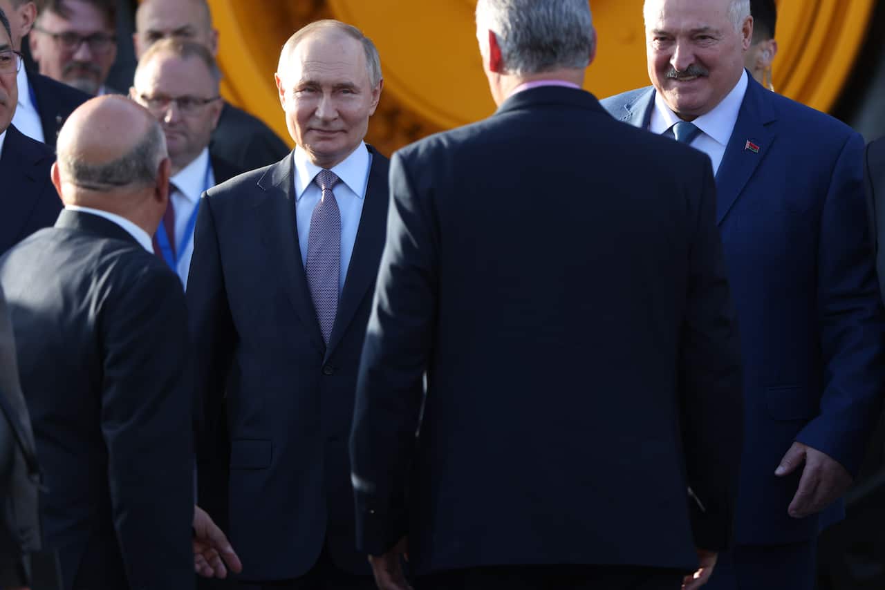 Russian President Vladimir Putin, wearing a dark suit and a patterned tie, stands among a group of men, including Belarusian President Alexander Lukashenko (right), who is smiling.