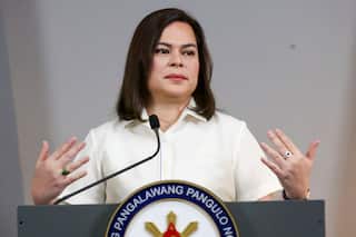 A woman with short brown hair and a white short sleeve button up shirt stands behind a podium. Her mouth is closed and both of her hands are raised to shoulder height, their backs facing the camera.