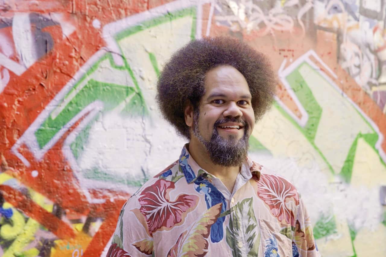 A man with an afro wearing a bright coloured shirt is smiling and standing in front of a grafitti covered wall