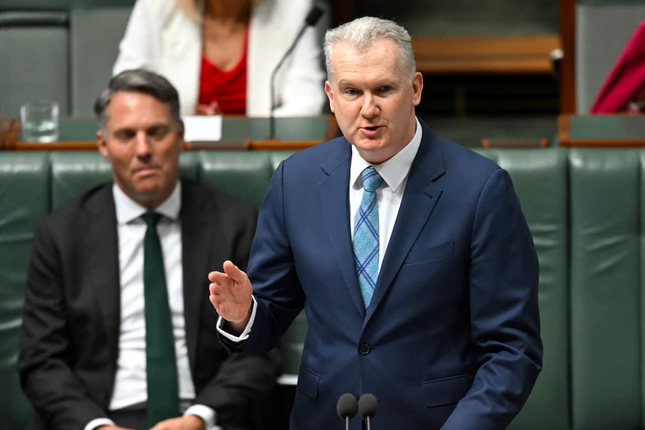 A man in a navy suit and light blue tie points a hand as he speaks.