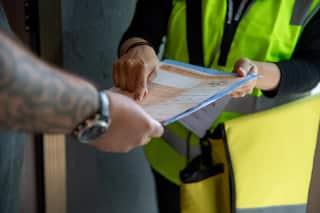 A woman wearing a fluorescent yellow vest hands a man an orange Census form. He wears a watch and has tattoos on his forearm.