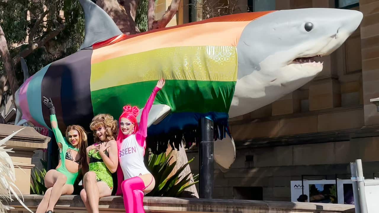 Three colourfully dressed performers posing in front of a rainbow-coloured shark model