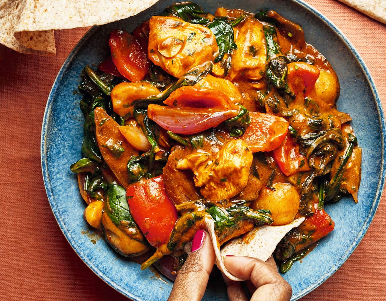 A colourful chicken and vegetable dish sits in a blue bowl. A hand can be seen scooping up some food with a folded piece of flatbread. 