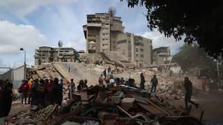 Rescue workers stand around a collapsed building with standing buildings behind it.