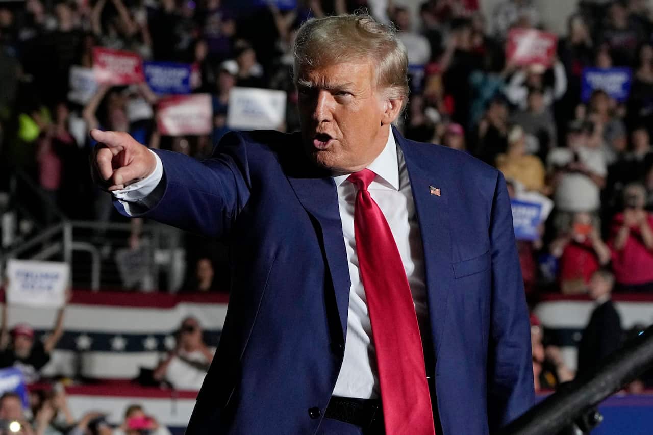 Donald Trump wearing a dark blue suit and red tie points forward with his index finger while speaking at a rally. 
