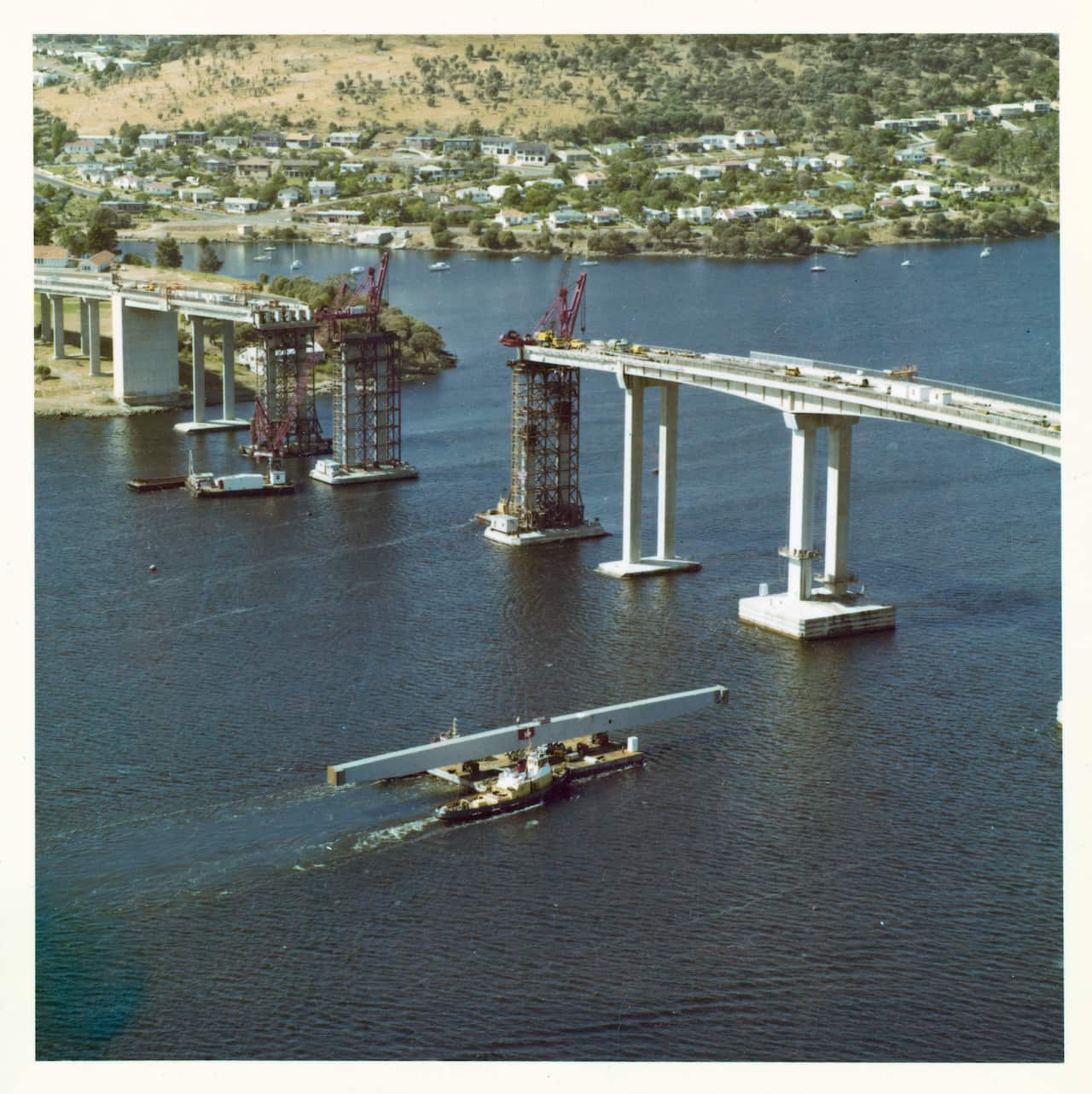 A river ferry transports a large concrete pylon towards a large bridge that is being repaired. 