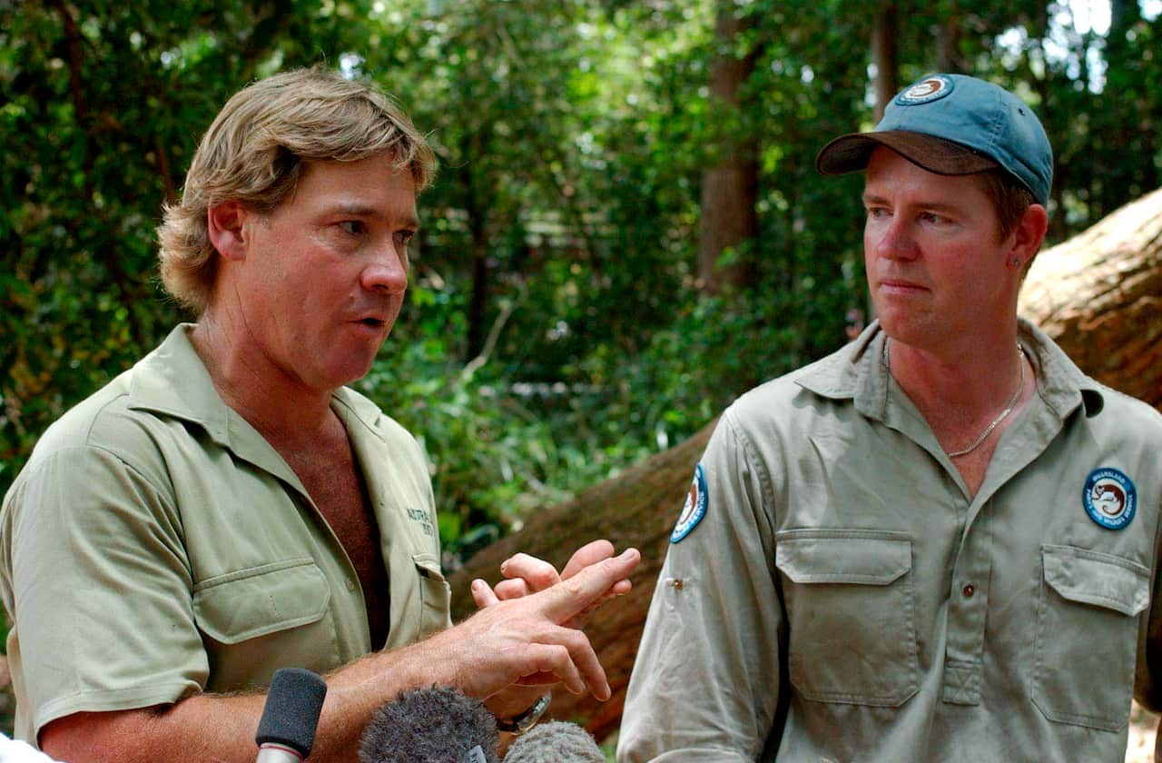 Two white men standing next to each other, both wearing green khaki shirts. A forest is behind them.