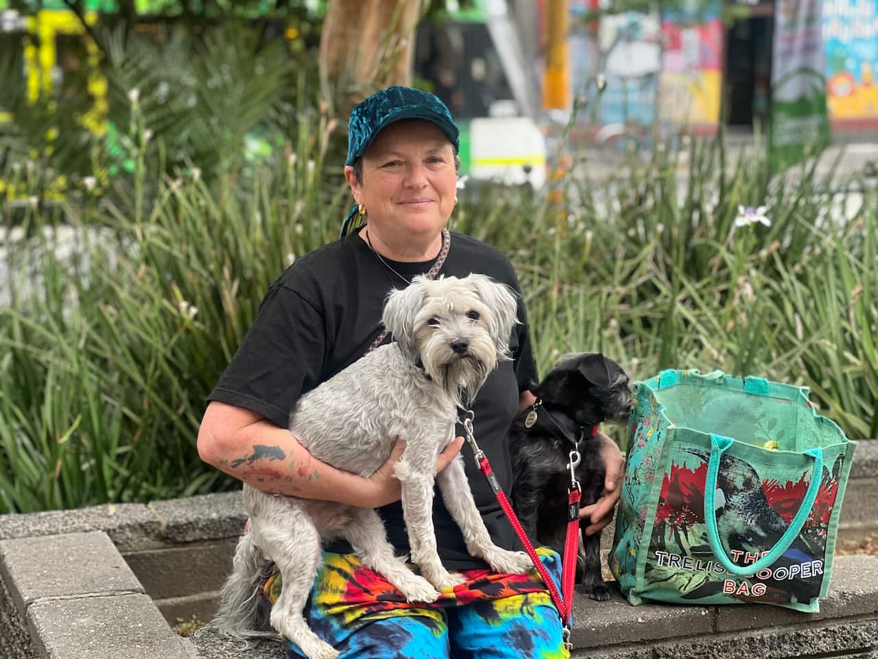 A woman in a blue cap, black T-shirt, and tie-dye trousers sits with a grey dog and a black dog.