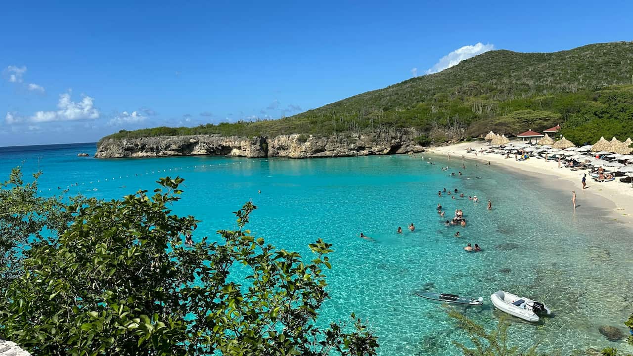 A beach with bright turqoie water and tents on the sand. The beach is surrounded by rocks and green forest. People are swimming and a boat is visible/ 