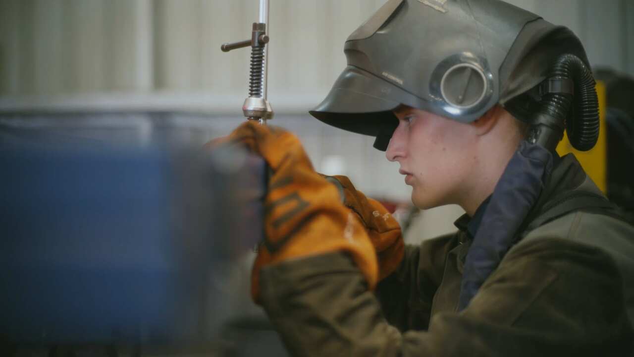 A young man is wearing protective gloves and a protective helmet as he focuses on a blue board which he assume is used for metal working 