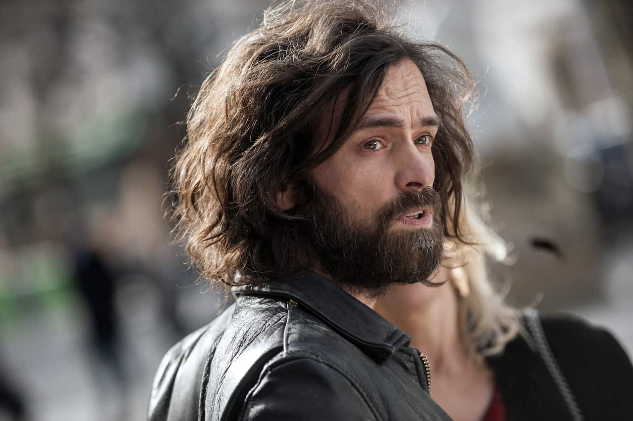 A man with a beard and chin-length wavy hair, wearing a black jacket, looks sideways.