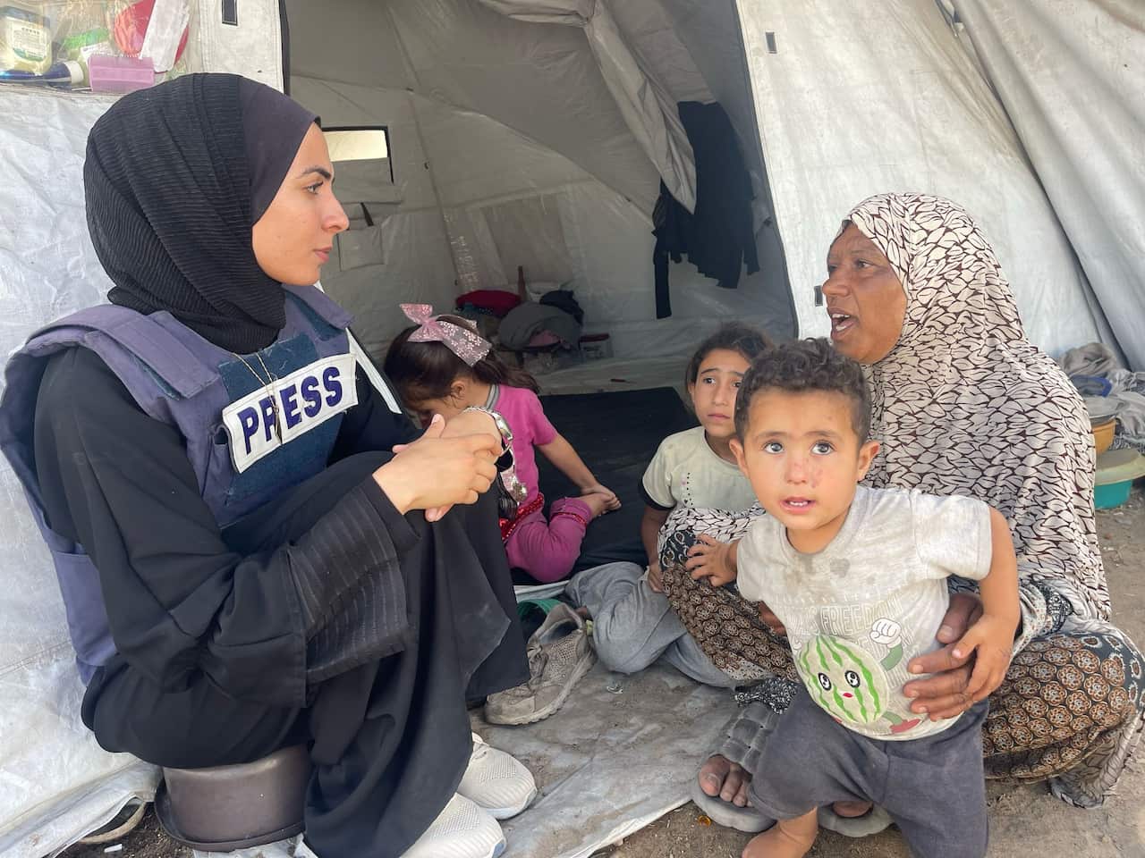 A young woman wearing a black hijab and press vest sits in front of a white tent talking to a woman and several small children.