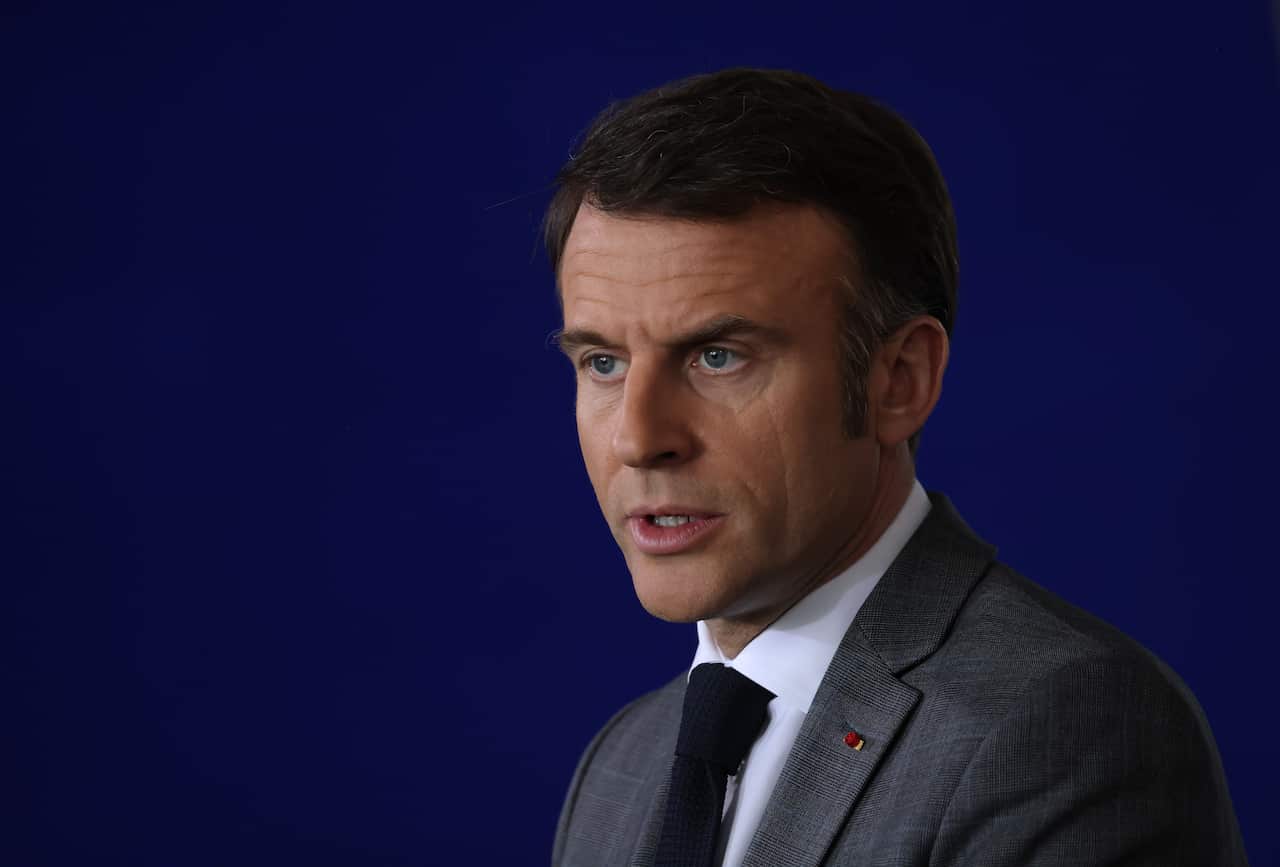 Emmanuel Macron, wearing a grey blazer, in front of a blue background.